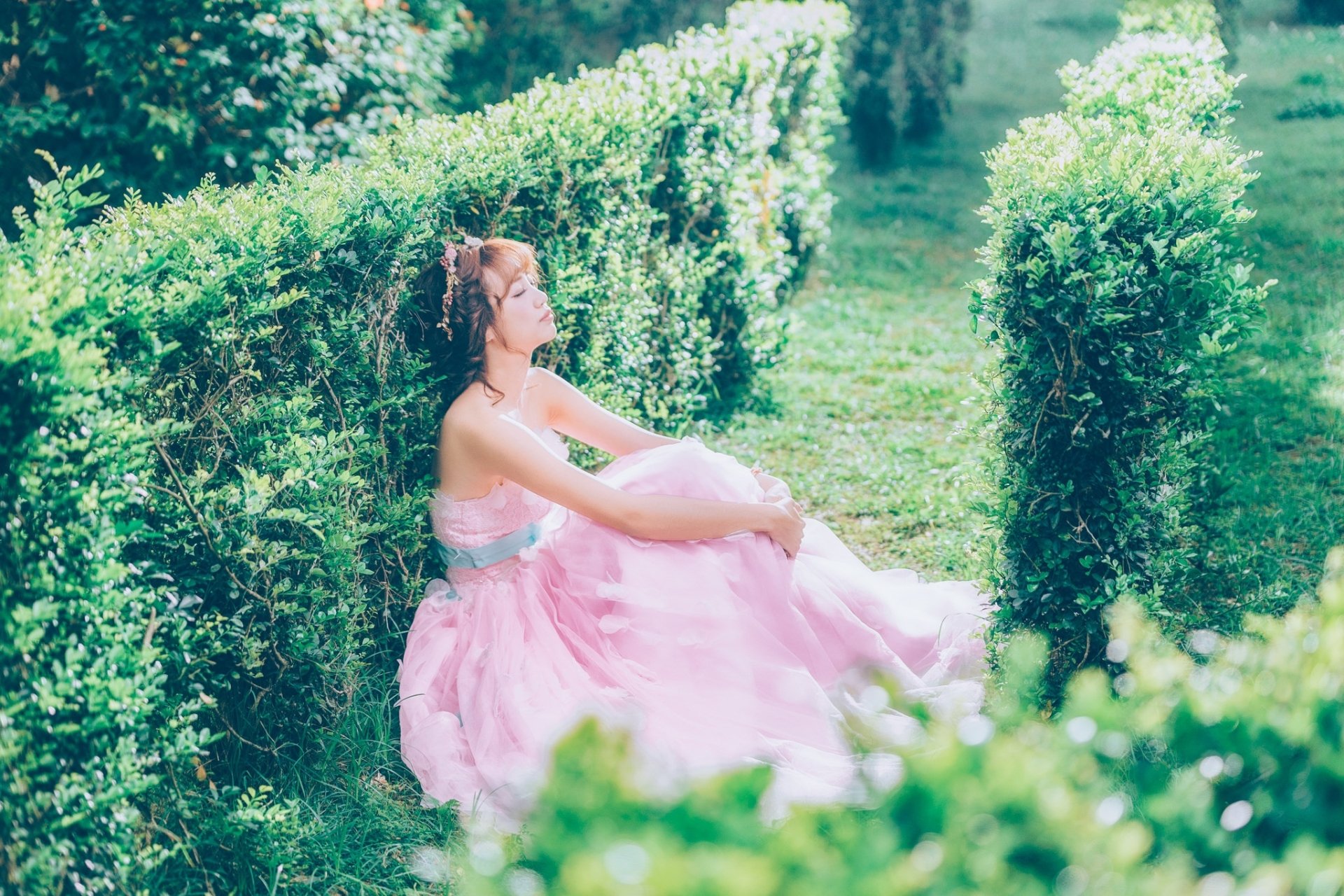 Sunlit HD PC desktop wallpaper of a woman model in a flowing pink dress seated among manicured green hedges, conveying a serene, dreamy mood.
