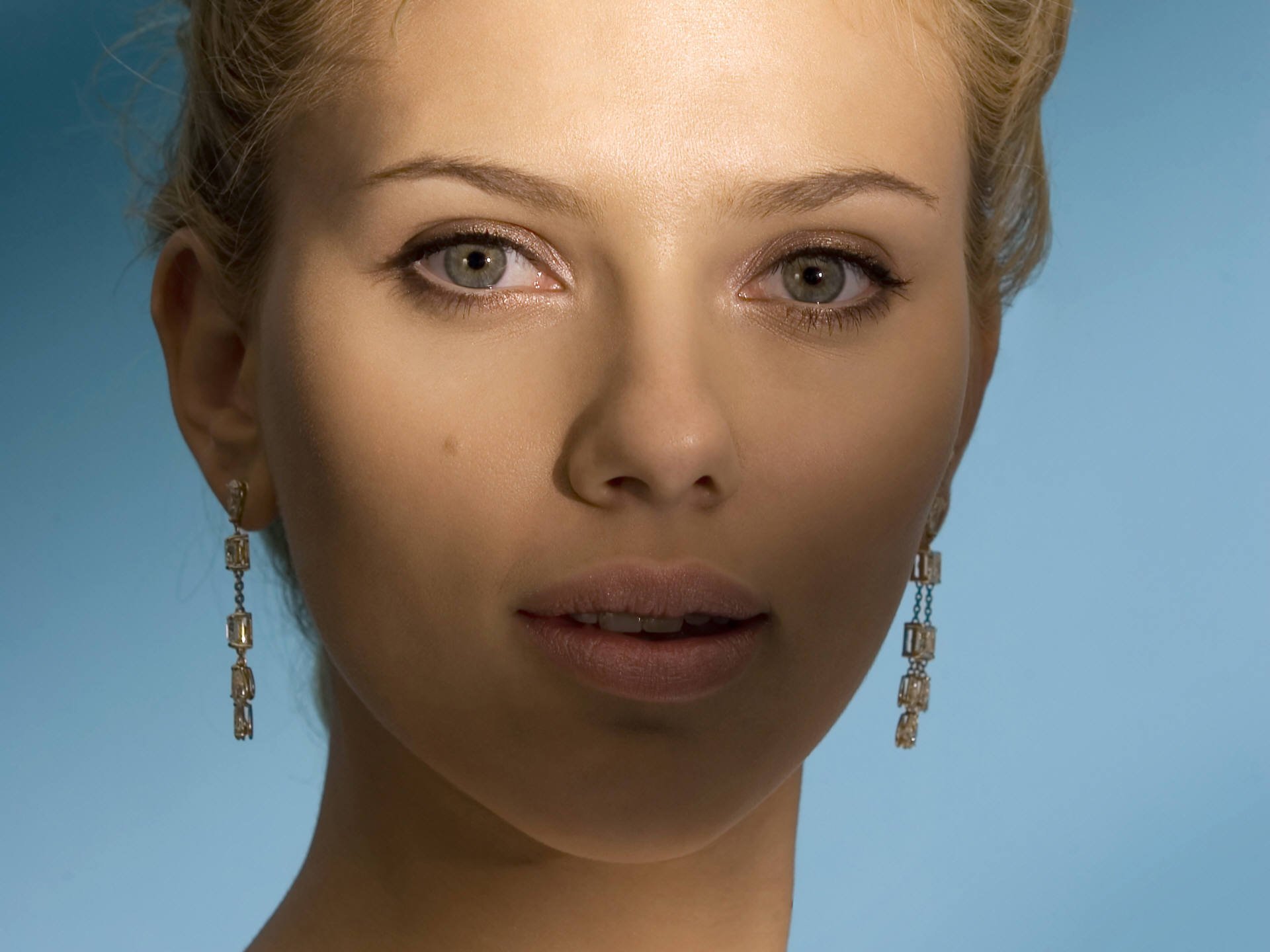 Close-up portrait of Scarlett Johansson with striking eyes and elegant earrings, set against a soft blue background. This HD image serves as a captivating desktop wallpaper.