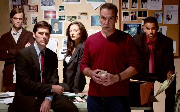 A high-definition wallpaper featuring the main characters of the TV show Criminal Minds, standing together around a cluttered desk in a tense investigative atmosphere.