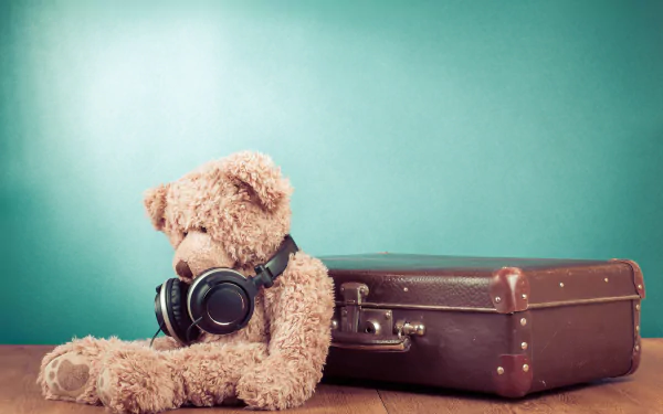 A fluffy teddy bear wearing headphones sits beside a vintage suitcase, set against a vibrant teal background, creating a cozy and nostalgic atmosphere. 