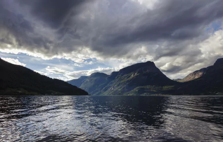 A serene view of Vangsmjøse lake in Norway, showcasing calm waters, dramatic mountains, and a cloudy sky. This stunning landscape makes for an inspiring HD desktop wallpaper.