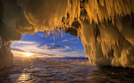 HD desktop wallpaper showcasing a winter ice cave with icicles hanging from the ceiling, framed by a vibrant sunset over a frozen landscape.