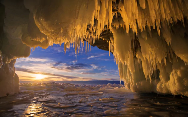 HD desktop wallpaper showcasing a winter ice cave with icicles hanging from the ceiling, framed by a vibrant sunset over a frozen landscape.