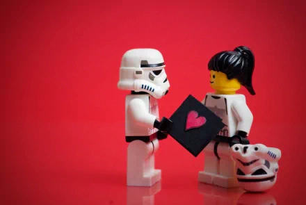 A creative HD desktop wallpaper featuring LEGO figures of a Stormtrooper offering a card with a heart to another LEGO figure holding a Stormtrooper helmet, set against a bright red background.