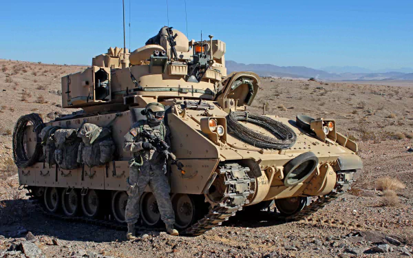 HD desktop wallpaper featuring a soldier standing beside an M2 Bradley armored personnel carrier in a desert military setting.