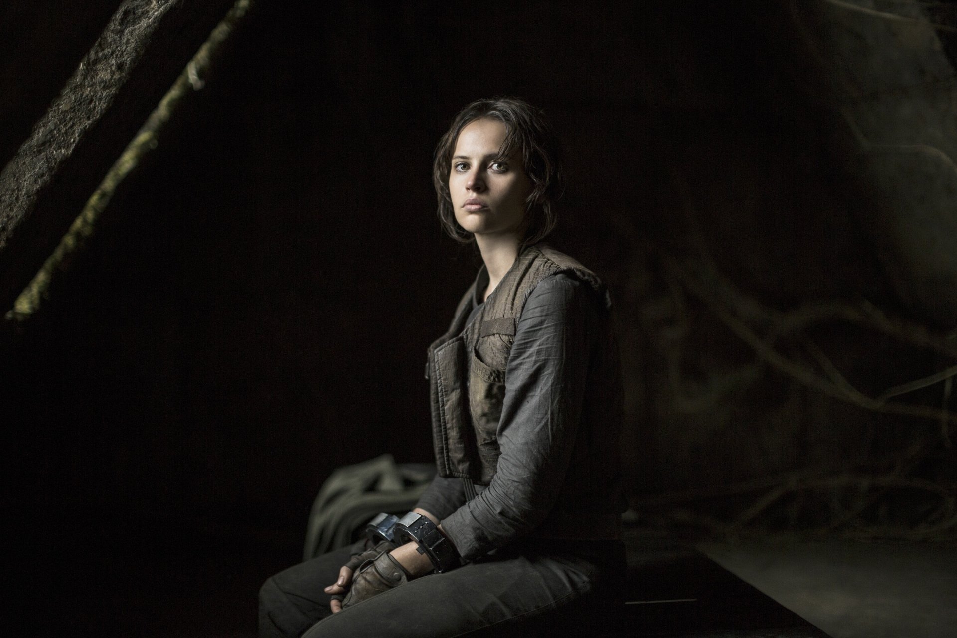 Jyn Erso, portrayed by Felicity Jones, sits contemplatively in a dark setting, capturing a moment from Rogue One: A Star Wars Story in this stunning 4K Ultra HD wallpaper.