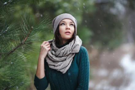 HD desktop wallpaper of model Angelina Petrova with blue eyes, wearing a gray hat and scarf, standing outdoors near pine branches in a snowy setting.