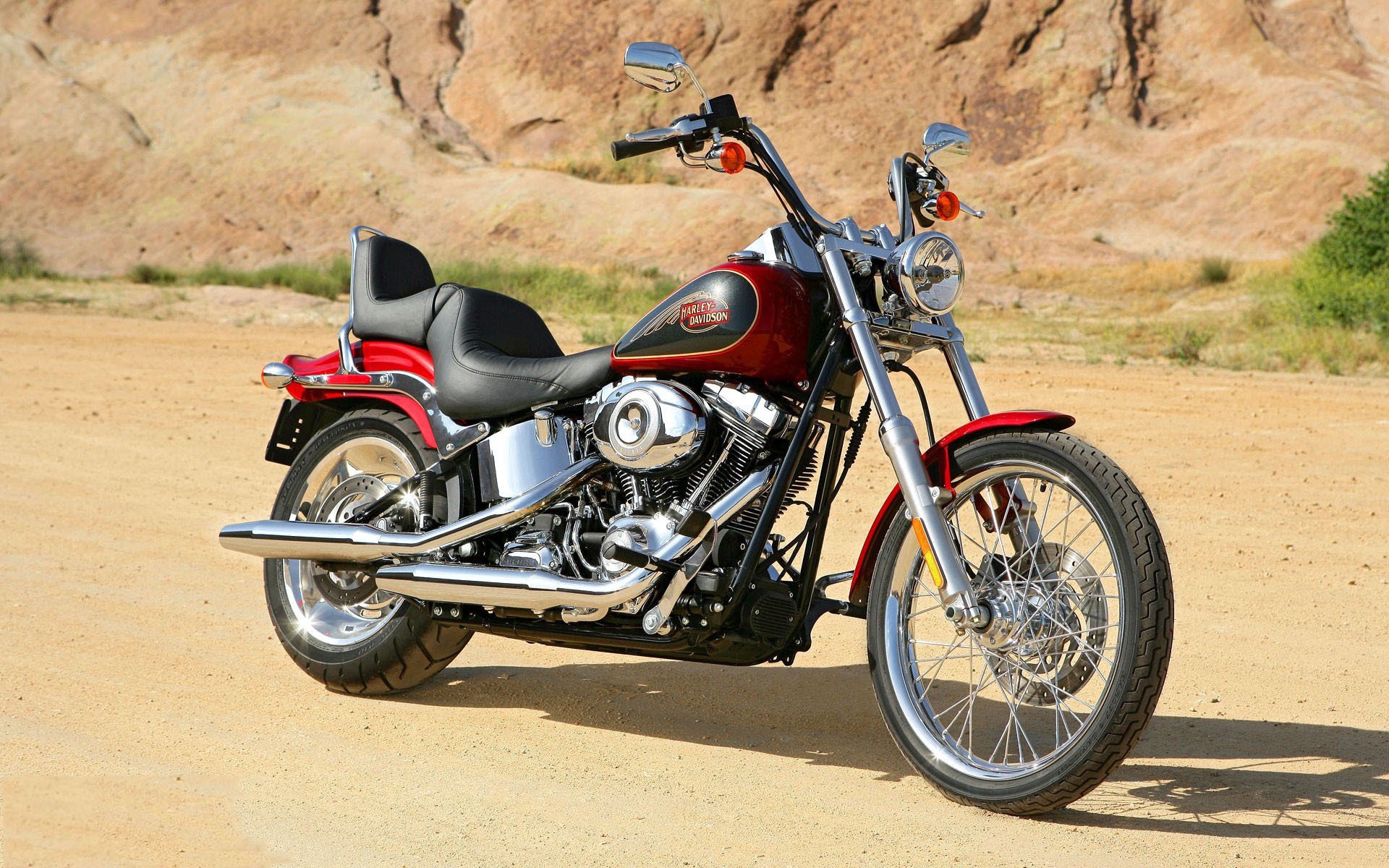 A red Harley-Davidson motorcycle parked on a sandy desert road with rocky hills in the background, featured as an HD PC desktop wallpaper.