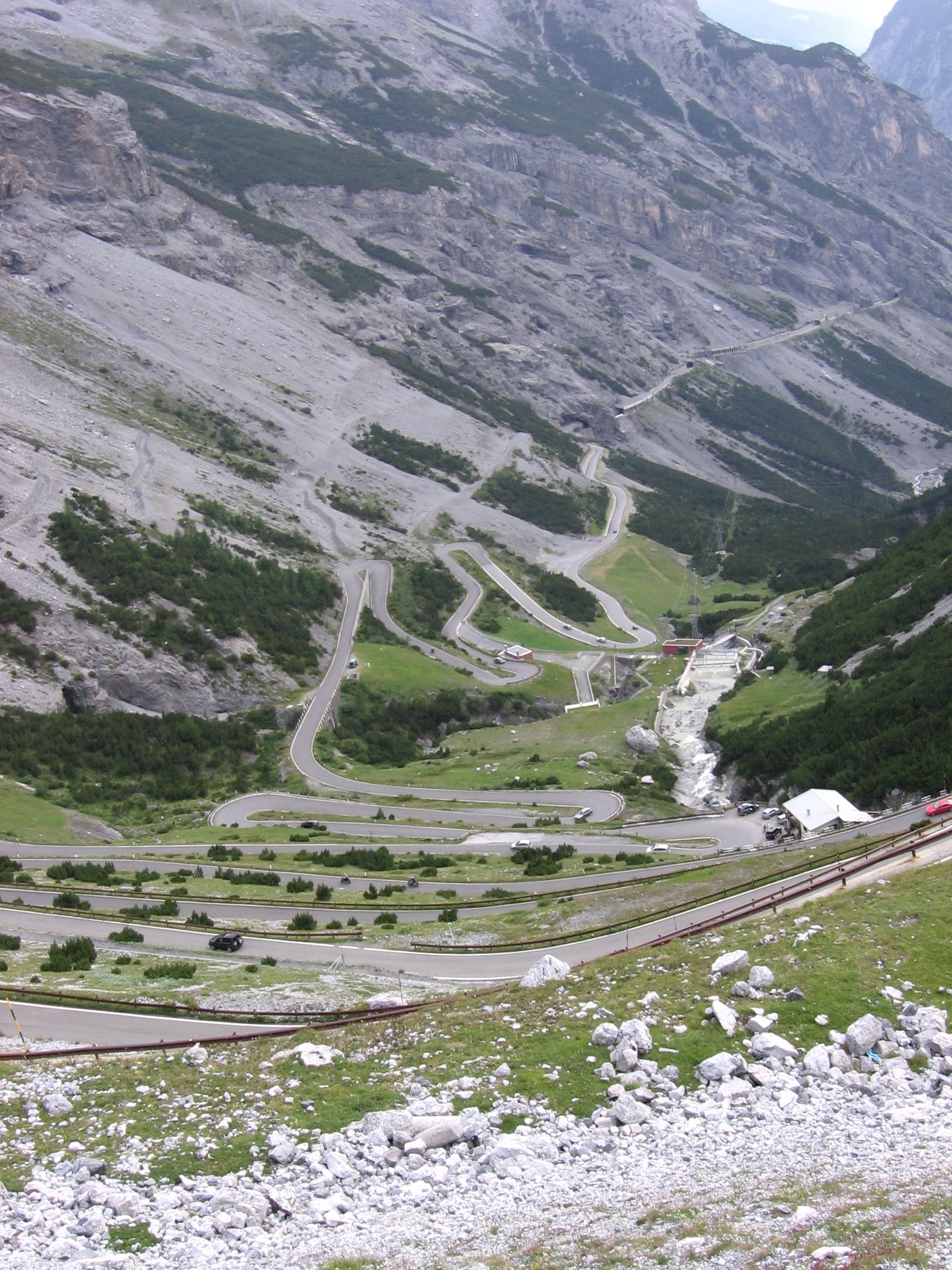 HD PC desktop wallpaper: nature landscape of a steep mountain with a winding switchback road descending into a green valley, a cascading stream and scattered alpine buildings.