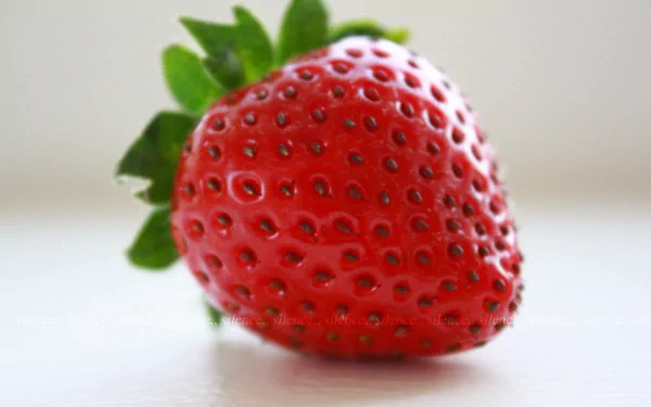 A vibrant, detailed strawberry sits atop a light surface, showcasing its rich red color and green leaves. This 4K Ultra HD image serves as an appealing desktop wallpaper.