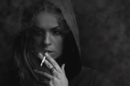 Black and white HD desktop wallpaper of a woman smoking, wearing a hooded jacket. The image captures a moody and contemplative atmosphere.