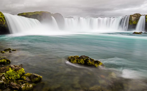4K Ultra HD wallpaper of a serene natural waterfall with white foam cascading into a clear turquoise pool surrounded by mossy rocks under a cloudy sky.