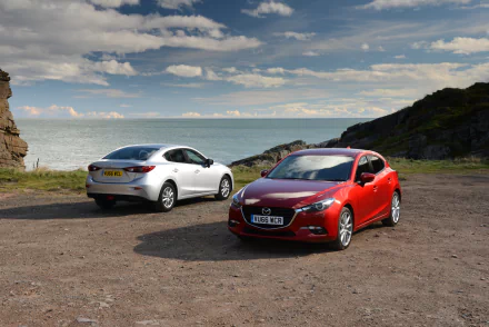 A white and a red Mazda 3 compact car parked on a coastal cliff under a partly cloudy sky, showcased in 4K Ultra HD desktop wallpaper quality.