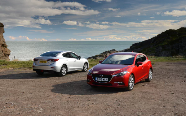A white and a red Mazda 3 compact car parked on a coastal cliff under a partly cloudy sky, showcased in 4K Ultra HD desktop wallpaper quality.