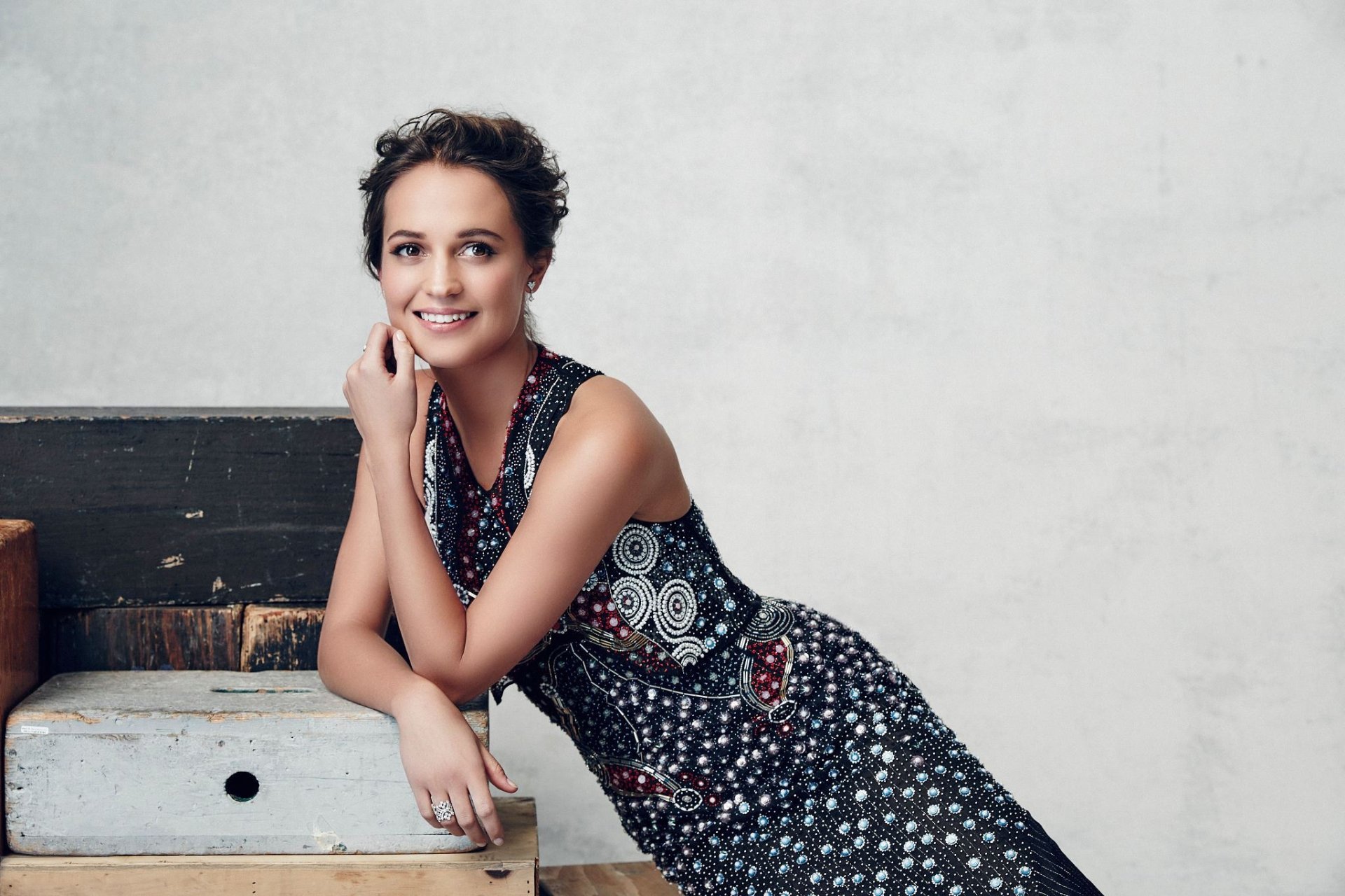 Alicia Vikander, Swedish actress with brunette hair and brown eyes, smiles while wearing a patterned dress against a neutral background in this HD desktop wallpaper.