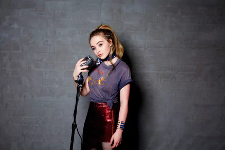 HD wallpaper of American singer Sabrina Carpenter with blonde hair and blue eyes, holding a microphone against a grey background.