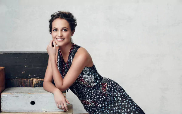 Alicia Vikander, Swedish actress with brunette hair and brown eyes, smiles while wearing a patterned dress against a neutral background in this HD desktop wallpaper.