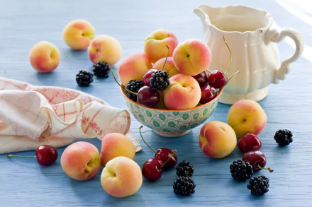 HD desktop wallpaper featuring a still life arrangement of fresh peaches, cherries, and blackberries on a blue surface with a white jug and cloth in the background.