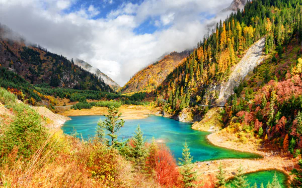 Vibrant landscape of Jiuzhaigou Park in China featuring a turquoise lake surrounded by colorful forested mountains under a partly cloudy sky.