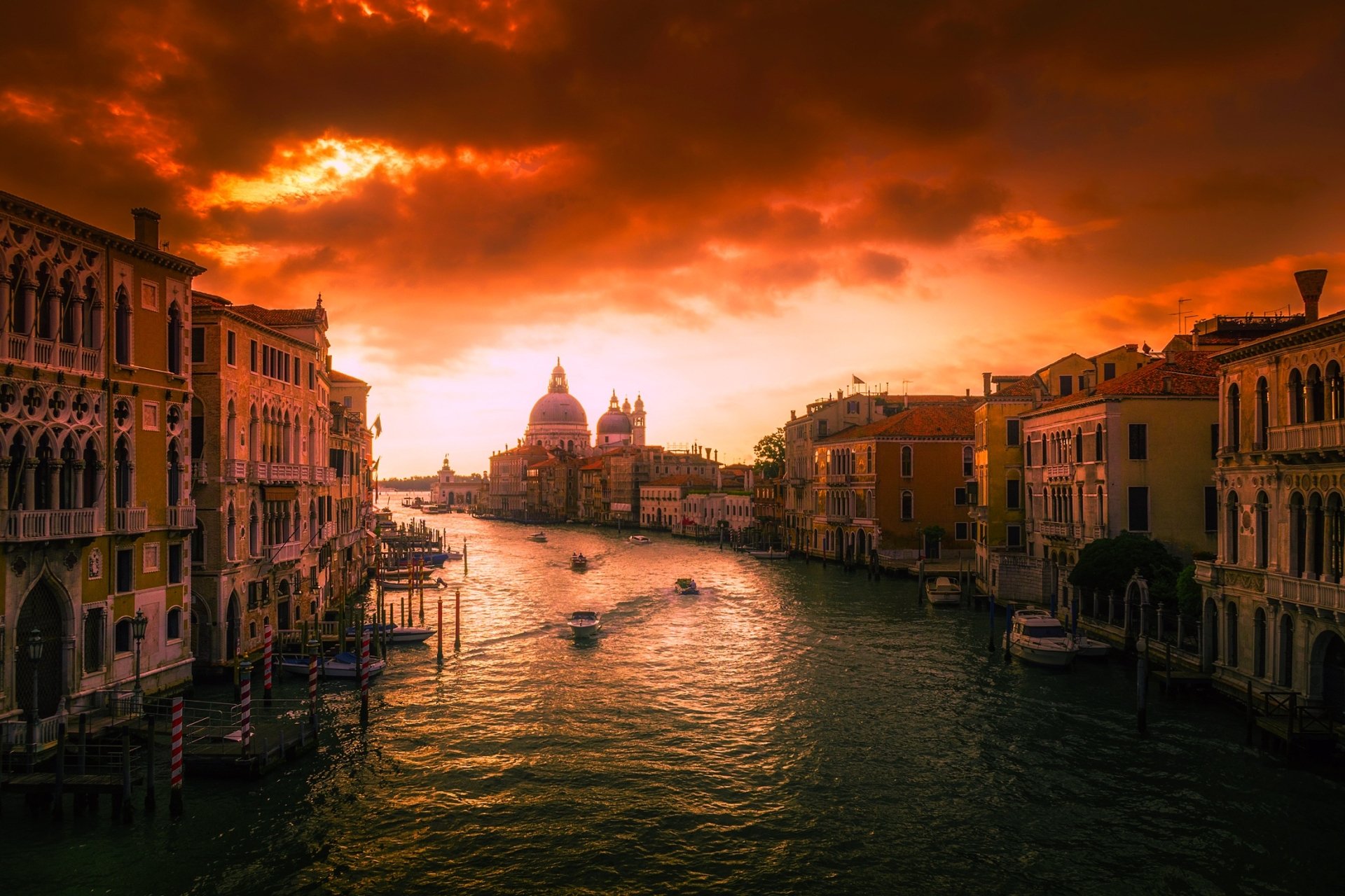 Sunset over the Grand Canal in Venice, Italy, with boats gliding through the water and historic buildings lining the cityscape under dramatic clouds.