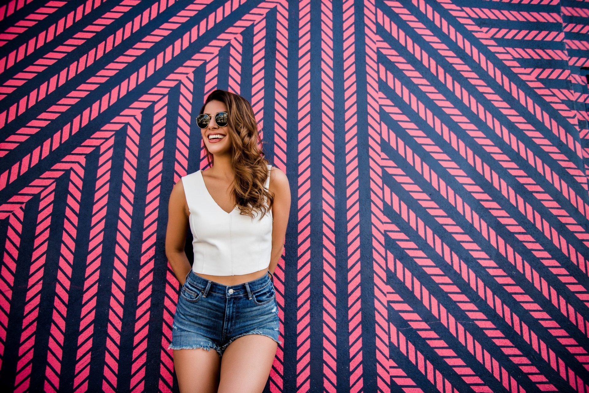 Brunette woman model with long hair and sunglasses smiling against a bold geometric pink-and-navy mural — HD PC desktop wallpaper background.