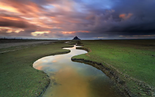 HD desktop wallpaper of Mont Saint-Michel in France, featuring a winding stream, dramatic clouds, and serene seascape at sunset with a religious landmark silhouette.