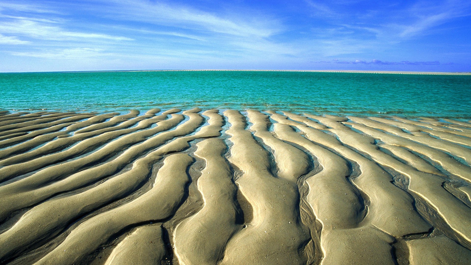 HD desktop wallpaper featuring a serene beach landscape, showcasing patterned sand dunes leading to vibrant turquoise waters under a clear blue sky.