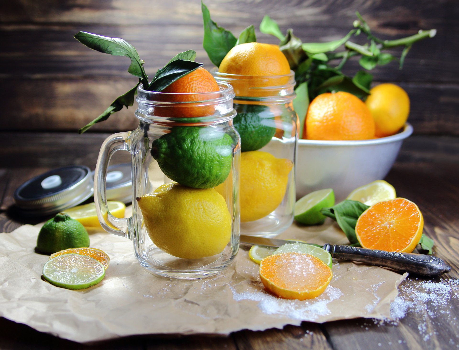 HD desktop wallpaper of a vibrant still life featuring fresh limes, lemons, and oranges arranged in glass jars and a bowl on a rustic wooden surface.