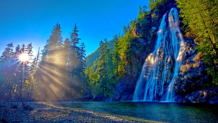 A stunning HD desktop wallpaper featuring a serene sunrise over a cascading waterfall, with sunbeams cutting through the trees, creating a peaceful nature scene.