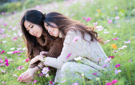 Two brunettes with brown eyes and warm smiles, posing in a flower-filled meadow. One is resting her head on the other's shoulder. The vibrant pink flowers add to the serene, intimate atmosphere.