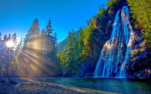 A stunning HD desktop wallpaper featuring a serene sunrise over a cascading waterfall, with sunbeams cutting through the trees, creating a peaceful nature scene.