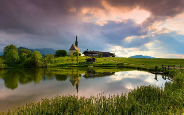 HD PC desktop wallpaper and background: serene mountain landscape with a small chapel on the shore of a reflective lake, grassy foreground, distant peaks and dramatic cloudy sky.