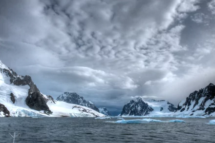 Snowy mountains and ice formations rise along the Lemaire Channel in Antarctica beneath a dramatic cloud-filled sky over the icy ocean waters.