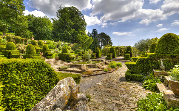 HD desktop wallpaper of a manicured garden featuring sculpted bushes, a stone fountain, and clear blue sky with scattered clouds, showcasing a serene man-made landscape.