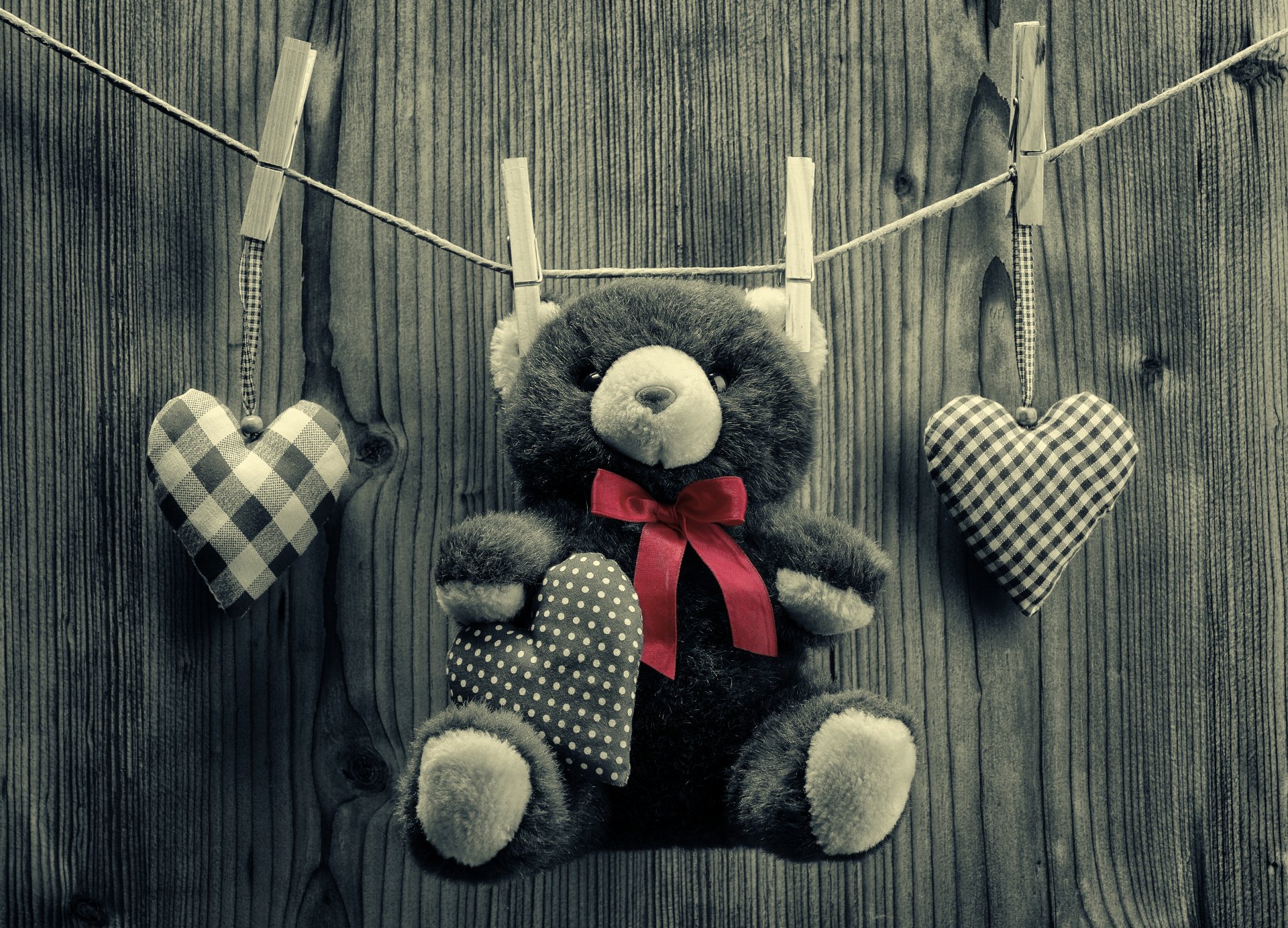 HD desktop wallpaper featuring a romantic stuffed teddy bear with a red bow tie, hanging on a clothesline with clothespins, accompanied by two gingham-patterned heart decorations against a wooden background.