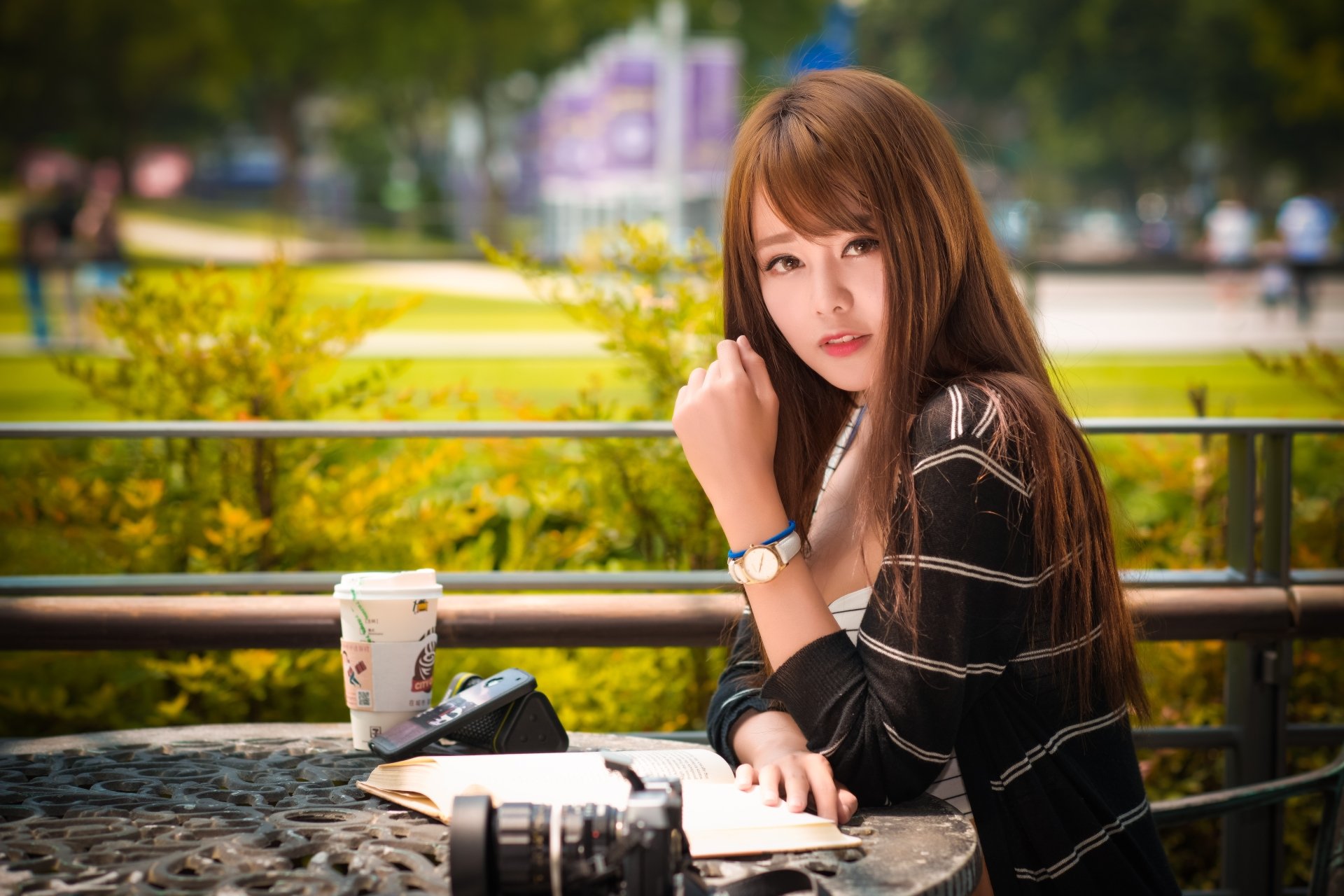 A brunette Asian woman poses outdoors for a high-definition wallpaper. She is sitting at a table with a book, a camera, and a cup, with a blurred, natural background providing depth of field.