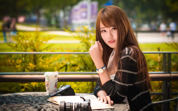 A brunette Asian woman poses outdoors for a high-definition wallpaper. She is sitting at a table with a book, a camera, and a cup, with a blurred, natural background providing depth of field.