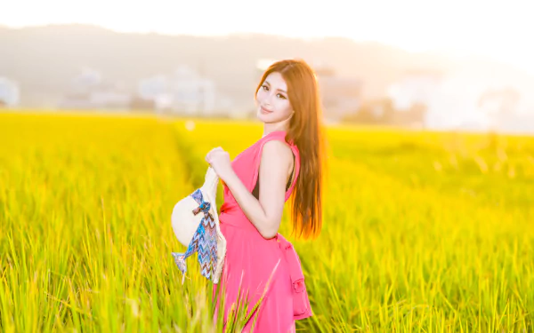 Asian woman with brunette hair wearing a pink dress and holding a hat stands in a sunlit field, captured with depth of field in vibrant 4K Ultra HD resolution.