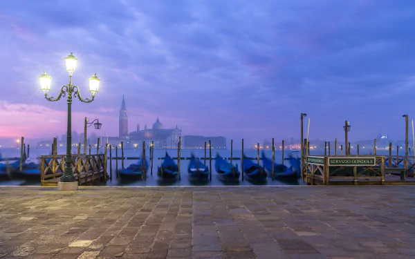 Dusk over a man-made dock in Venice, Italy, featuring gondolas lined up on calm water beneath a glowing street lamp.