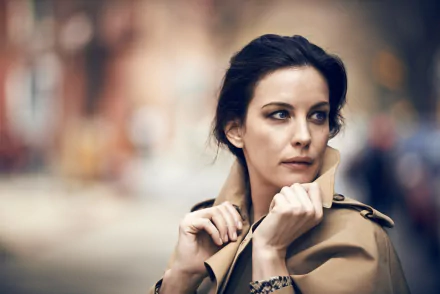 HD desktop wallpaper featuring actress and brunette celebrity Liv Tyler in a contemplative pose, wearing a beige coat against a blurred outdoor background.
