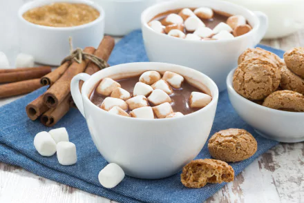 A cozy HD desktop wallpaper featuring white cups of hot chocolate topped with marshmallows, cinnamon sticks, and a bowl of cinnamon cookies on a blue cloth.