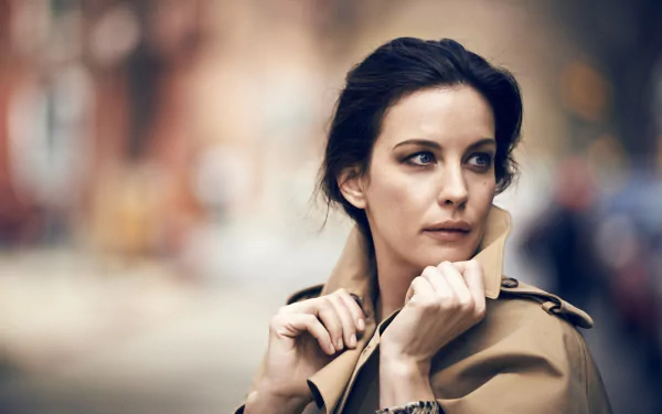 HD desktop wallpaper featuring actress and brunette celebrity Liv Tyler in a contemplative pose, wearing a beige coat against a blurred outdoor background.