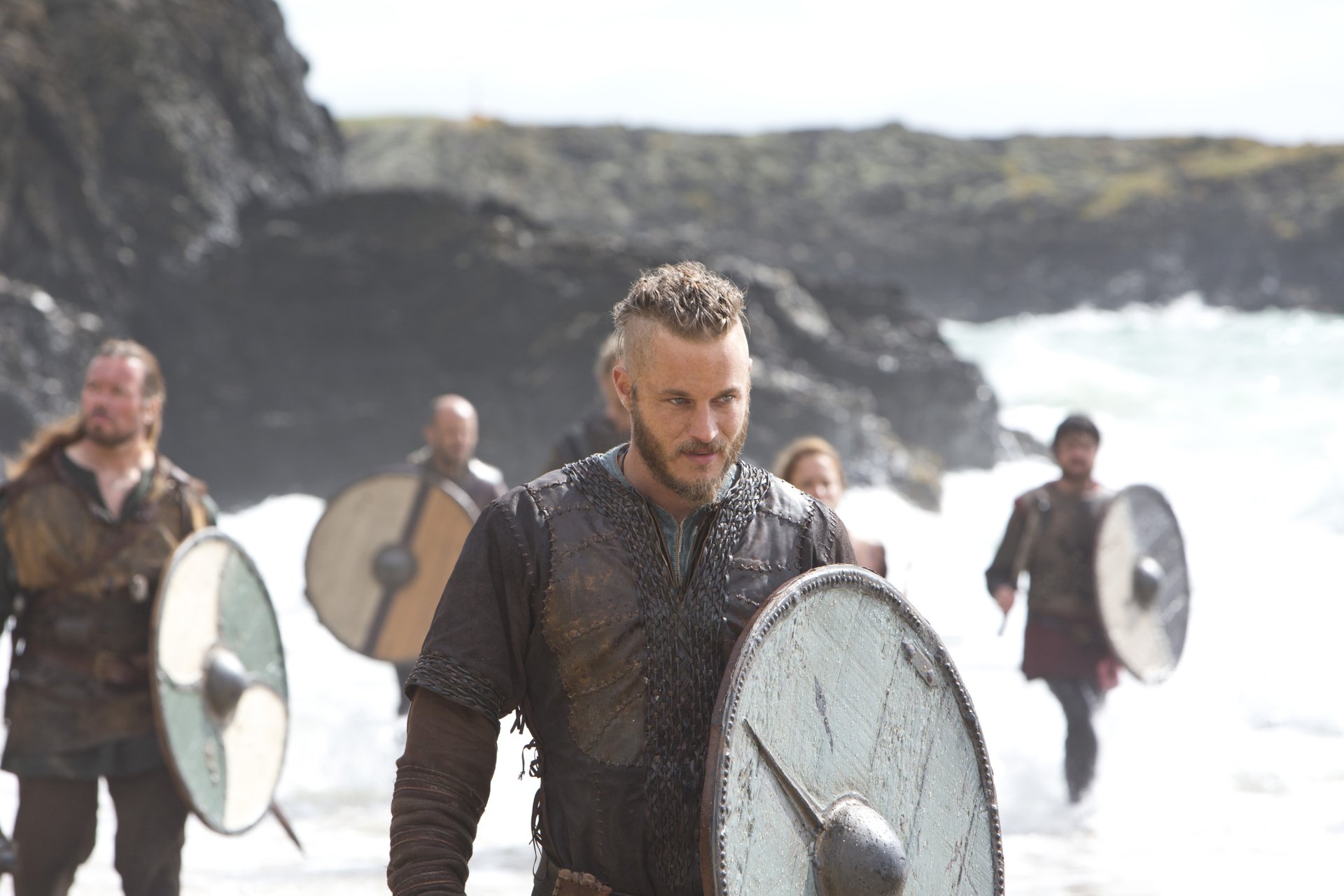 Vikings (2013) TV show 2K Quad HD PC wallpaper: bearded warrior in leather armor holding a round shield, leading shield-bearers along a windswept rocky shoreline.