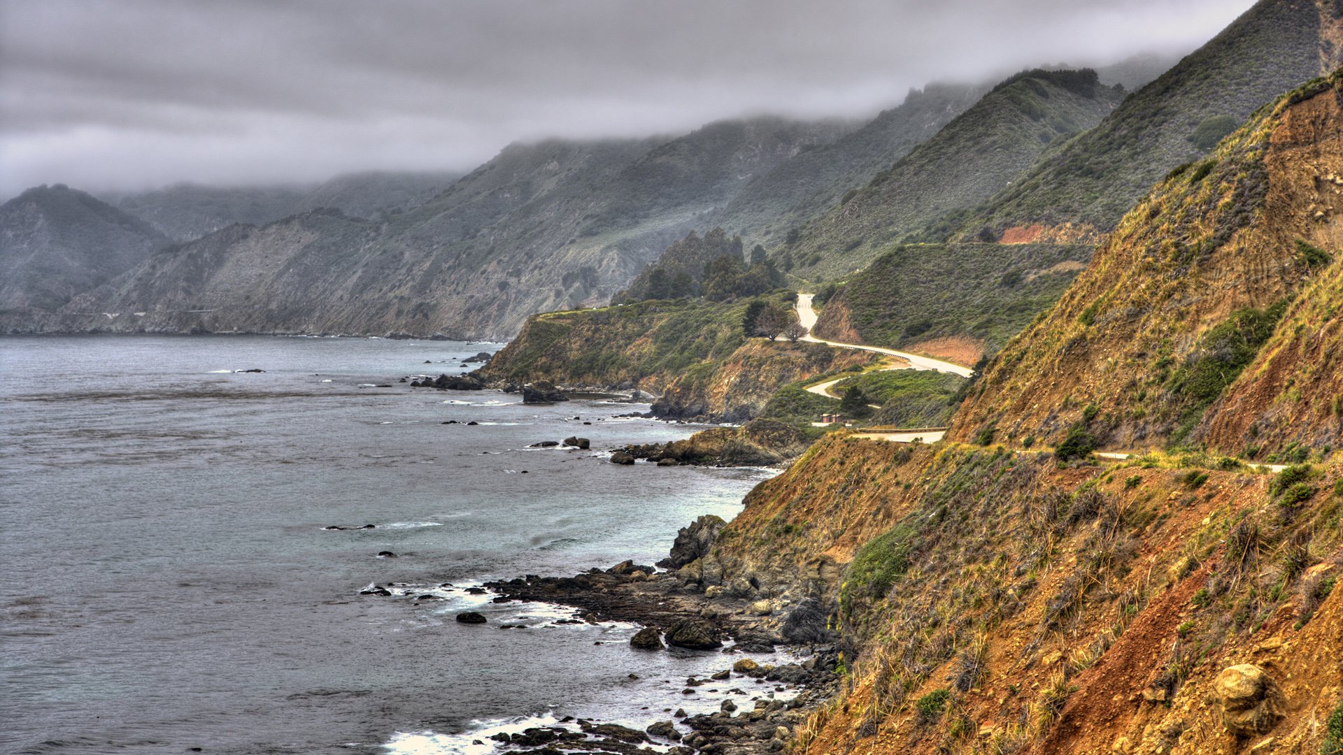 HD PC desktop wallpaper showing a rugged coastline with rocky cliffs, winding coastal road, and mist-covered mountains under an overcast sky.
