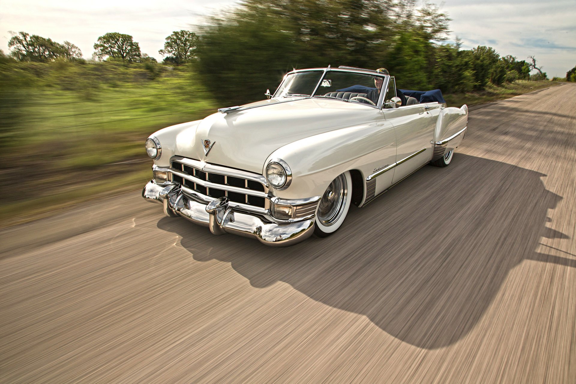 HD PC wallpaper of a white Cadillac Series 62 convertible vehicle — a lowrider luxury hot rod cruising a tree-lined road with motion blur.