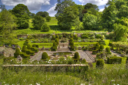 A beautifully landscaped garden featuring neatly trimmed bushes, a central fountain, and vibrant greenery under a clear blue sky. This high-definition wallpaper exudes tranquility and natural beauty.