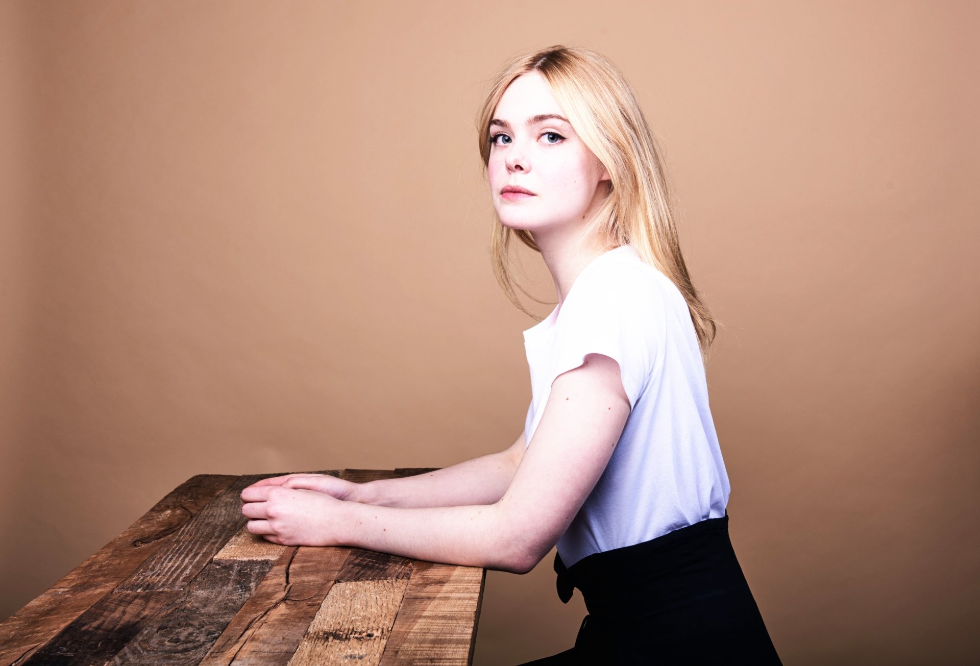 Elle Fanning, American actress with blonde hair and green eyes, poses against a plain beige background in this 8K Ultra HD desktop wallpaper.