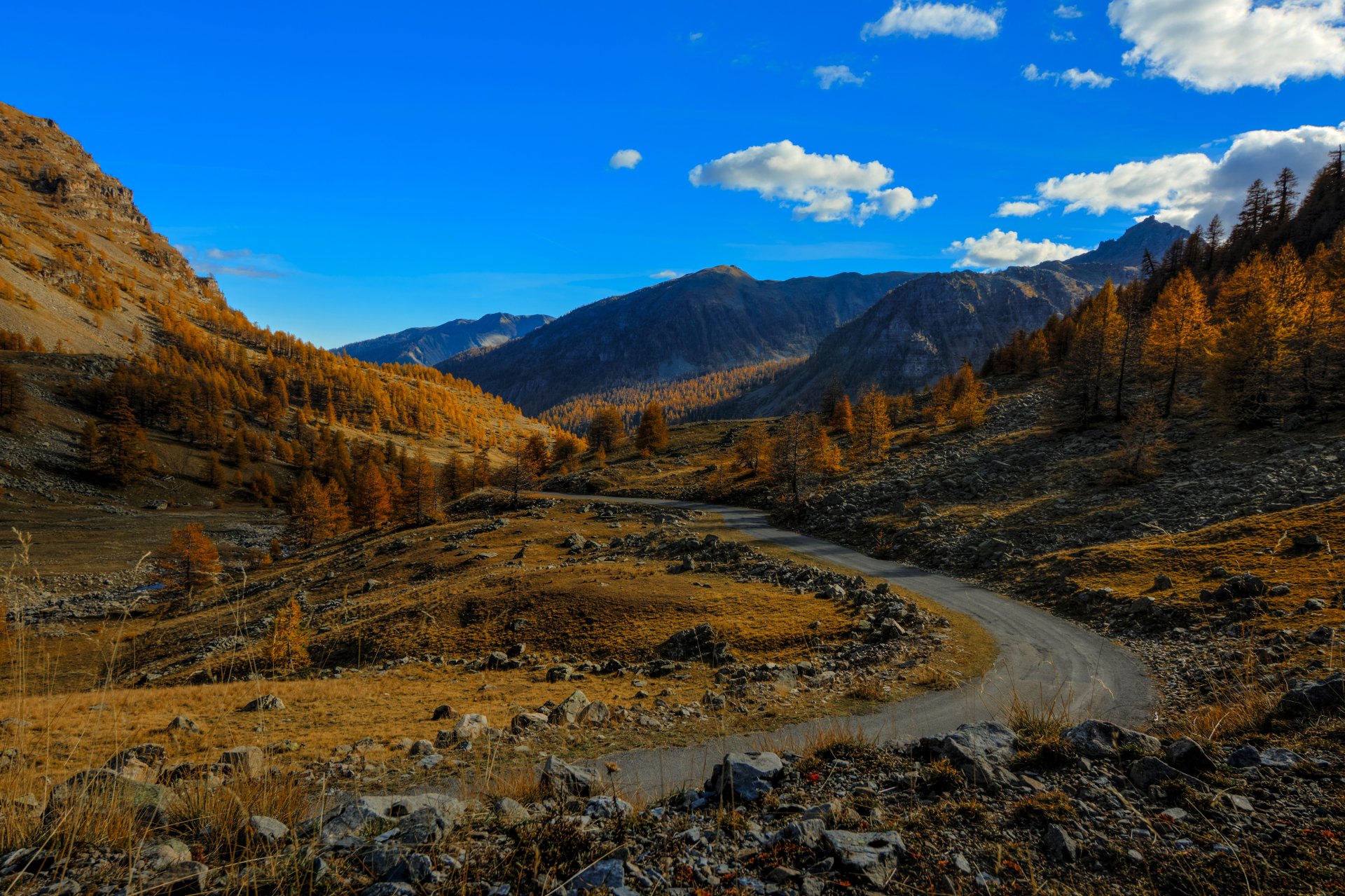 4K Ultra HD PC desktop wallpaper: winding dirt road through an autumn mountain valley, golden fall trees and rocky slopes beneath a vivid blue sky — nature landscape.