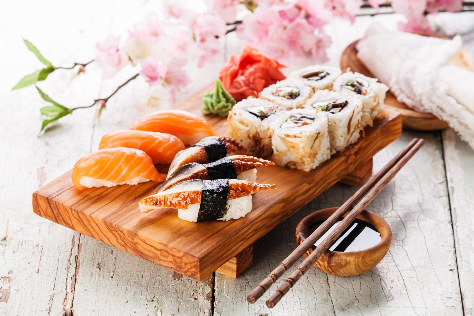 HD desktop wallpaper featuring a still life of assorted sushi, including rice, fish, and seafood, artfully arranged on a wooden board with chopsticks and cherry blossoms.