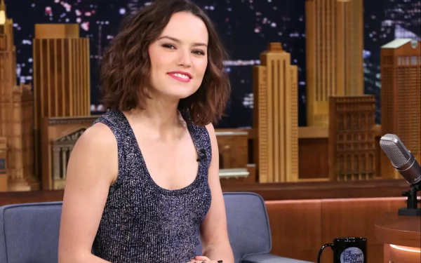Brunette actress and celebrity Daisy Ridley smiles while seated on a talk show set, featured in a high-definition PC desktop wallpaper and background.